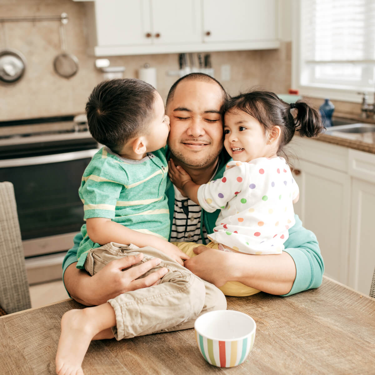 Dad in kitchen with kids sitting on table and giving him a hug